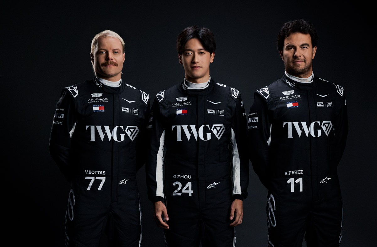 Three Team Members Wearing Matching Black Racing Suits With Cadillac And Sponsor Logos Stand Side By Side Against A Dark Studio Background.